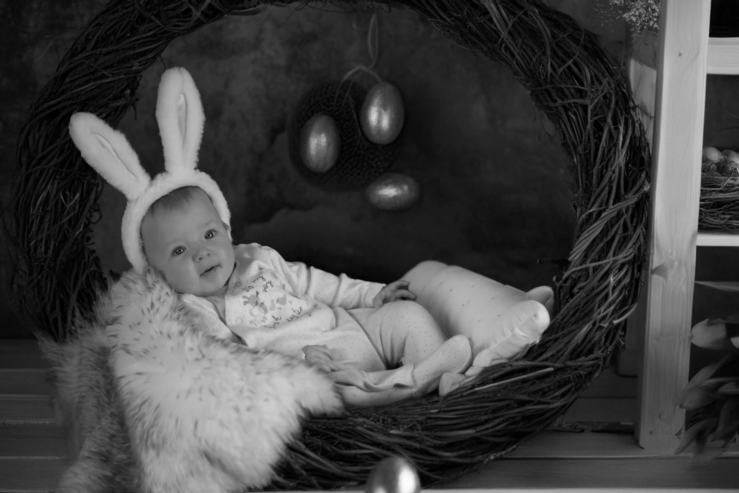Black and white photograph of a baby in a bunny costume lying in a decorative nest with Easter eggs.