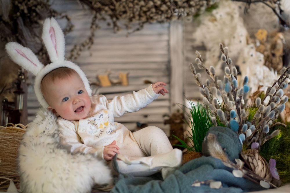 Baby wearing bunny ears sits on fuzzy white rug surrounded by spring floral decor and reaches upward with joy.