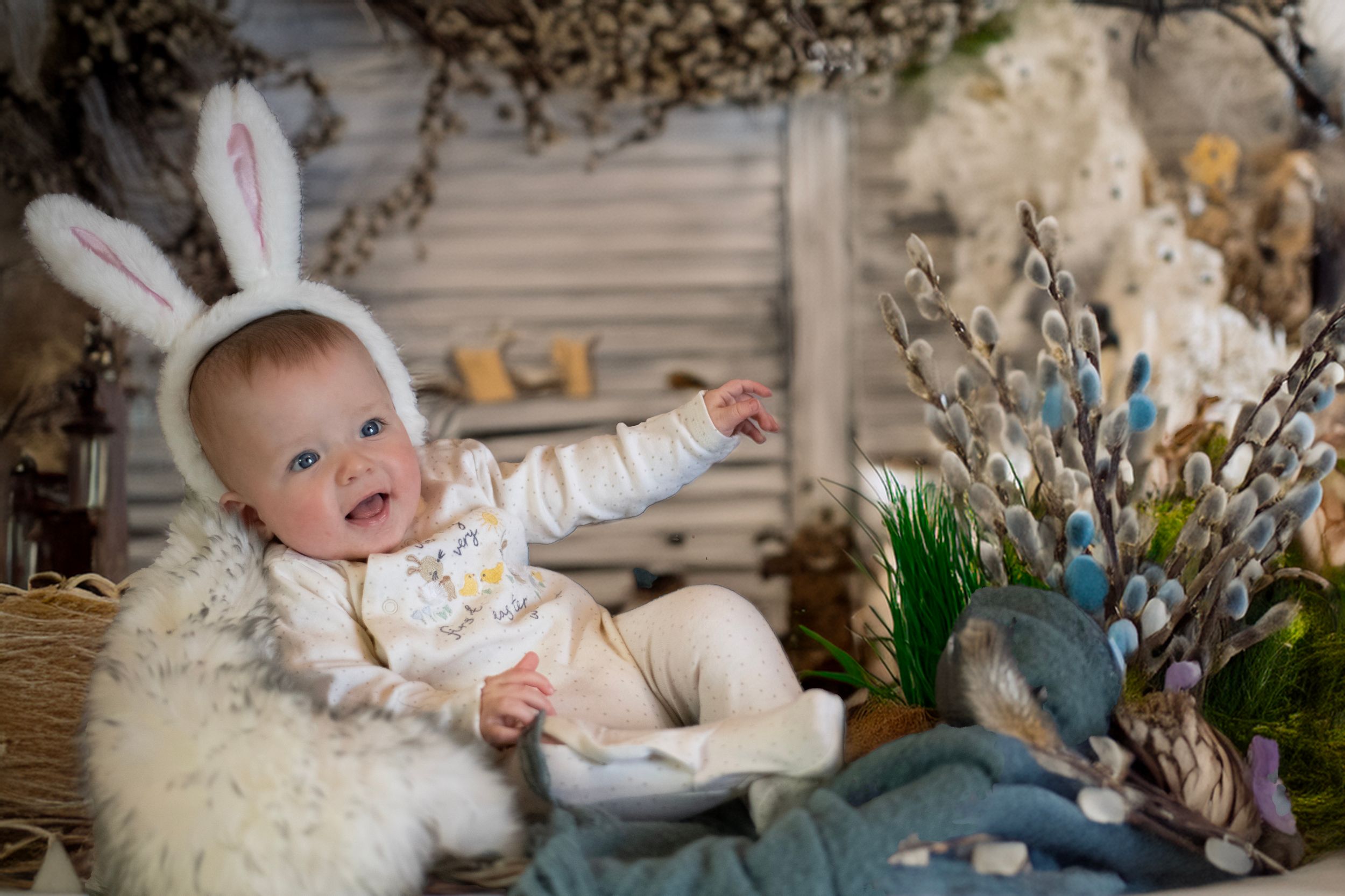 Baby wearing bunny ears sits on fuzzy white rug surrounded by spring floral decor and reaches upward with joy.
