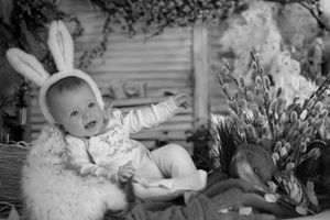 Black and white image of an infant wearing bunny ears sitting on a furry seat next to spring flowers.