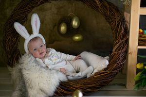 A small baby wearing bunny ears rests in a cozy Easter nest setting with golden eggs in the background.
