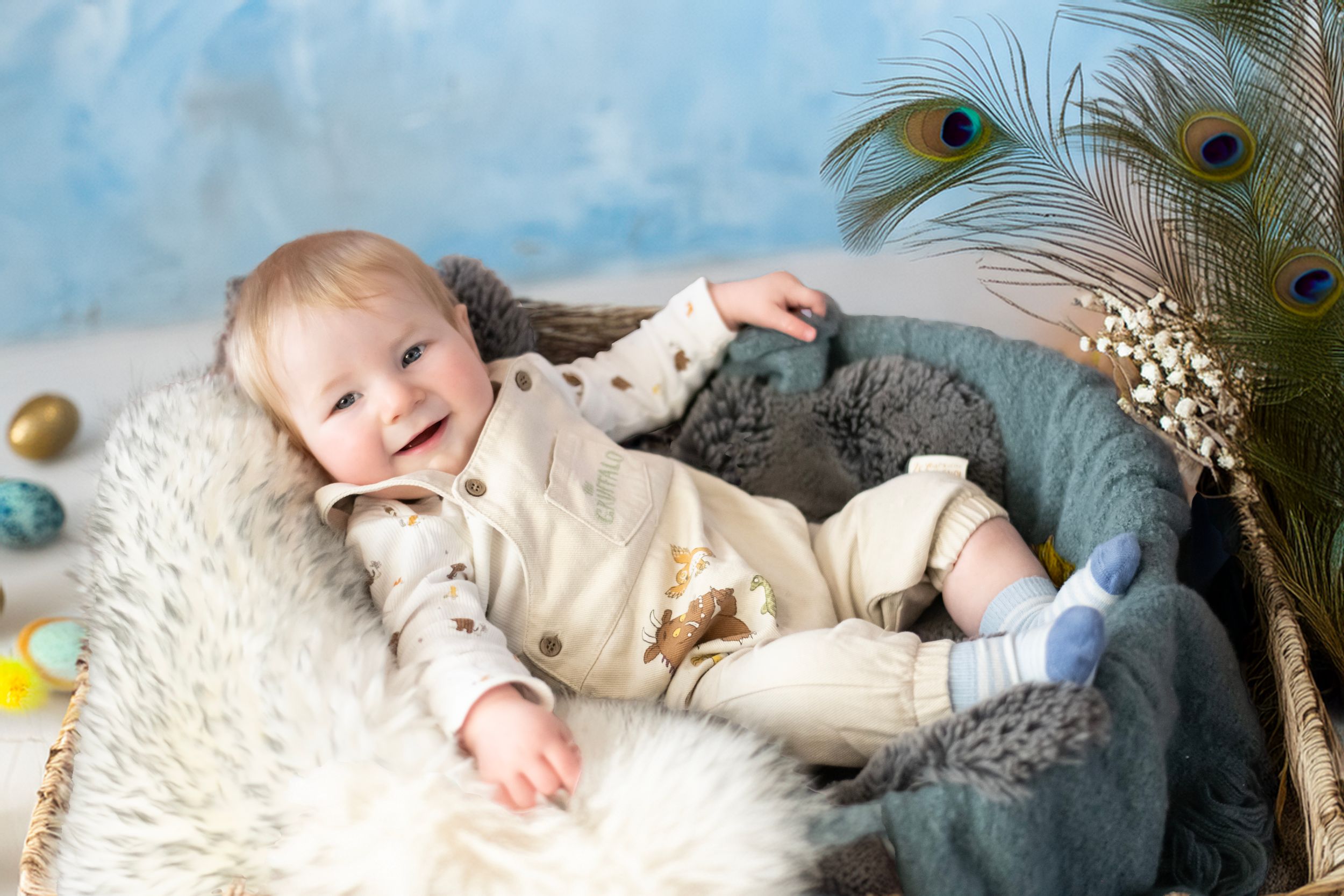 Baby lying on fluffy white blanket with peacock feather decoration and blue backdrop in cozy photo setting.
