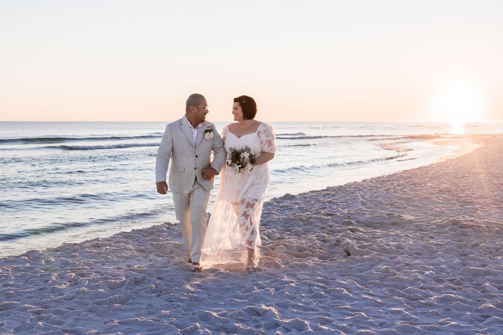 A couple in white attire shares a romantic moment on a sandy beach at sunset by the ocean waves.