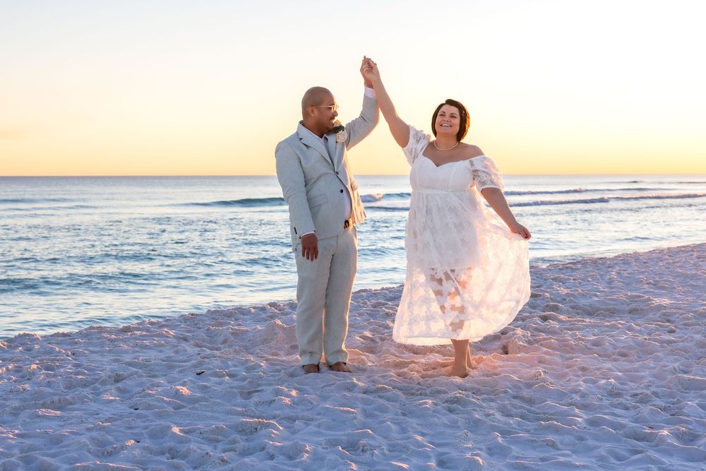 A couple in white attire dances joyfully on a sandy beach at sunset.