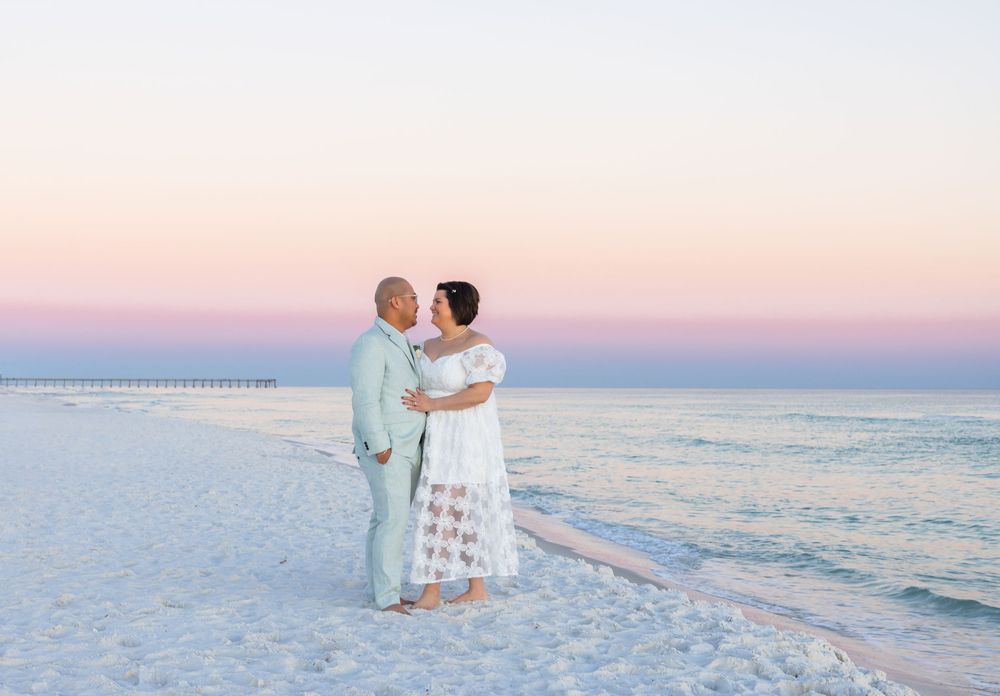 Couple in white outfits walking along pristine white sand beach at sunset with ocean waves in background.