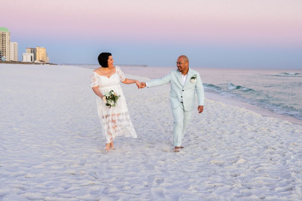 Couple in light blue and white outfits walking together on a pristine white sandy beach at sunset.