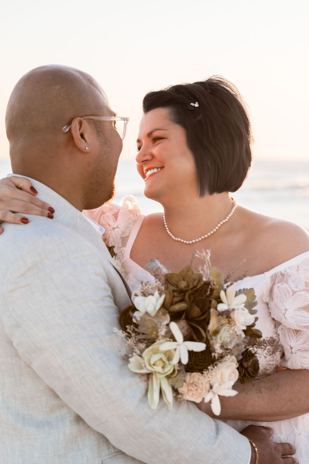 A romantic moment captured between a couple on the beach with white floral bouquet and elegant attire.