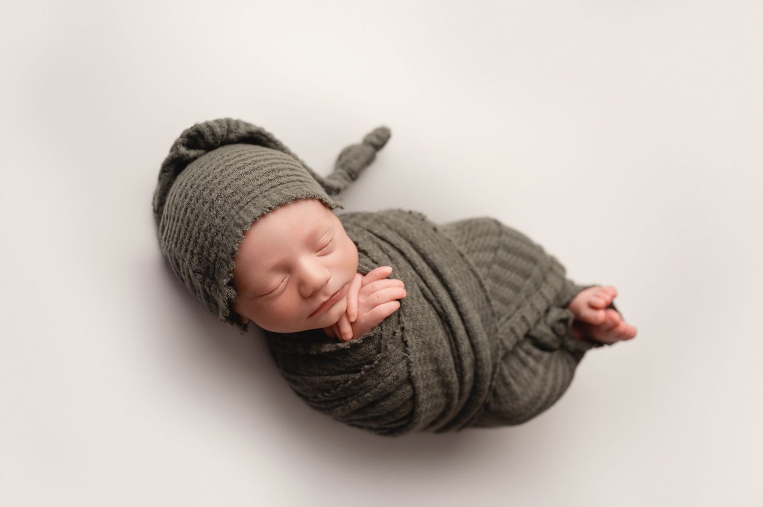 Upper St Clair Newborn baby wrapped in cozy green gray knit swaddle and matching hat sleeping peacefully on white background.