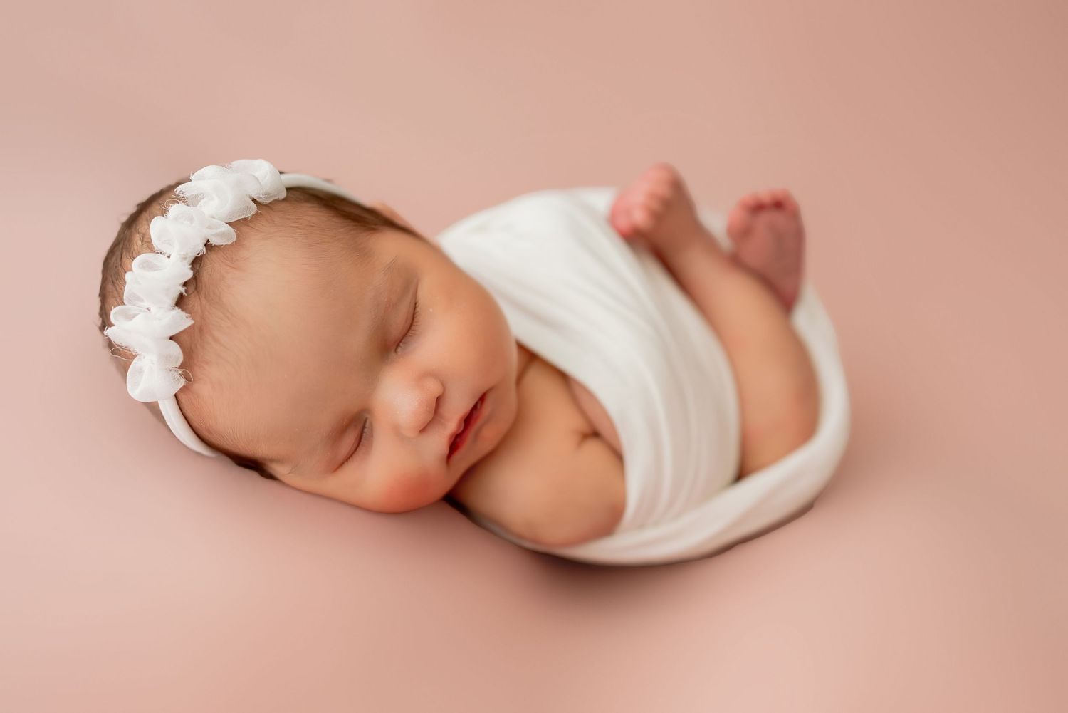 Peaceful sleeping newborn baby wrapped in white fabric wearing a delicate floral headband on pink background.
