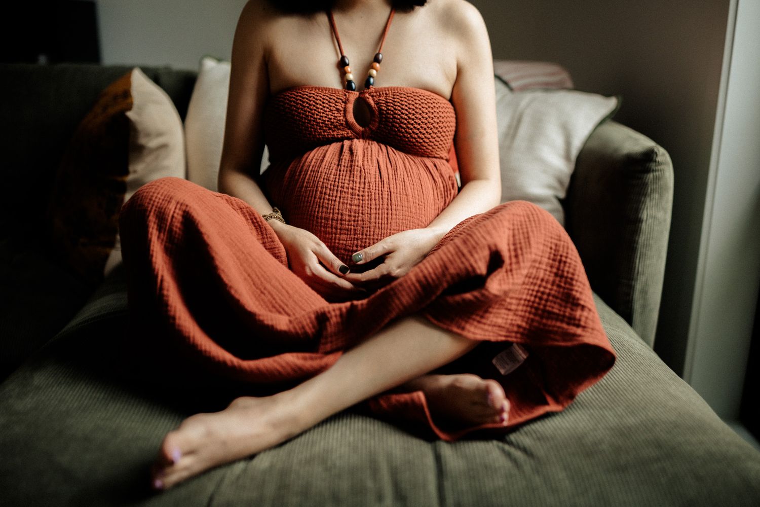 Pregnant person in a rust-colored dress sits cross-legged on grey couch in moody lighting.