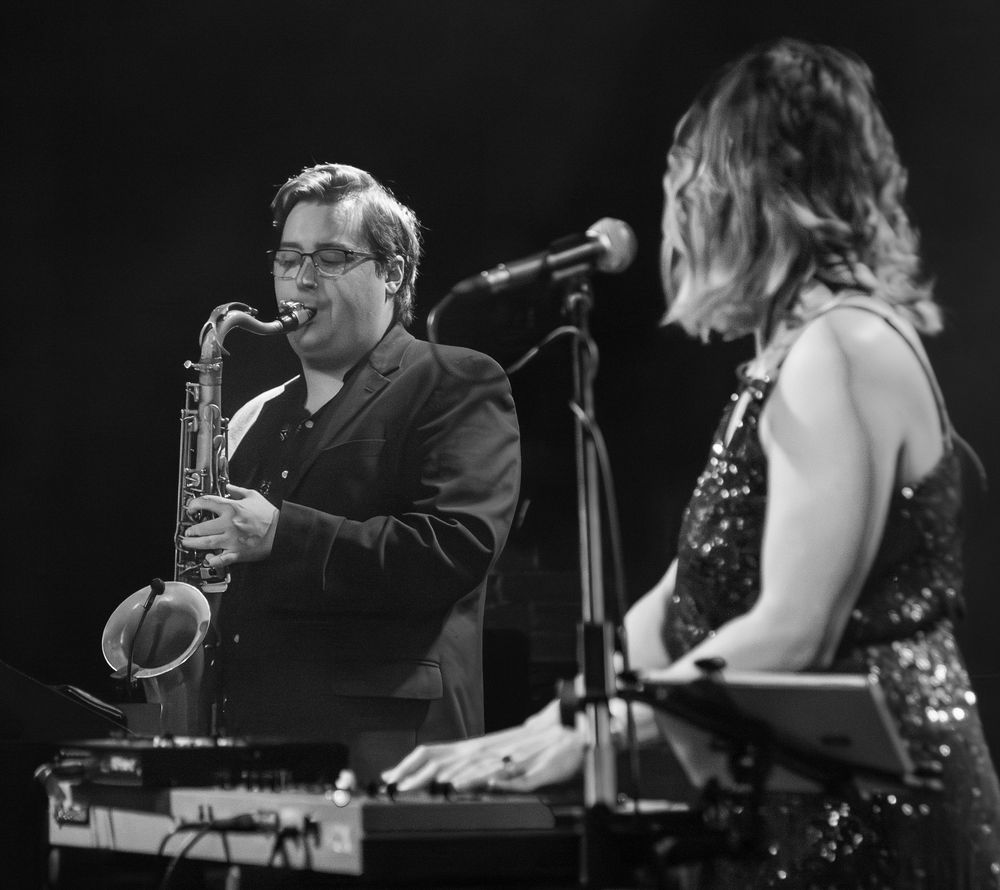 Man playing the saxophone with lead vocalist admiring him while playing piano.