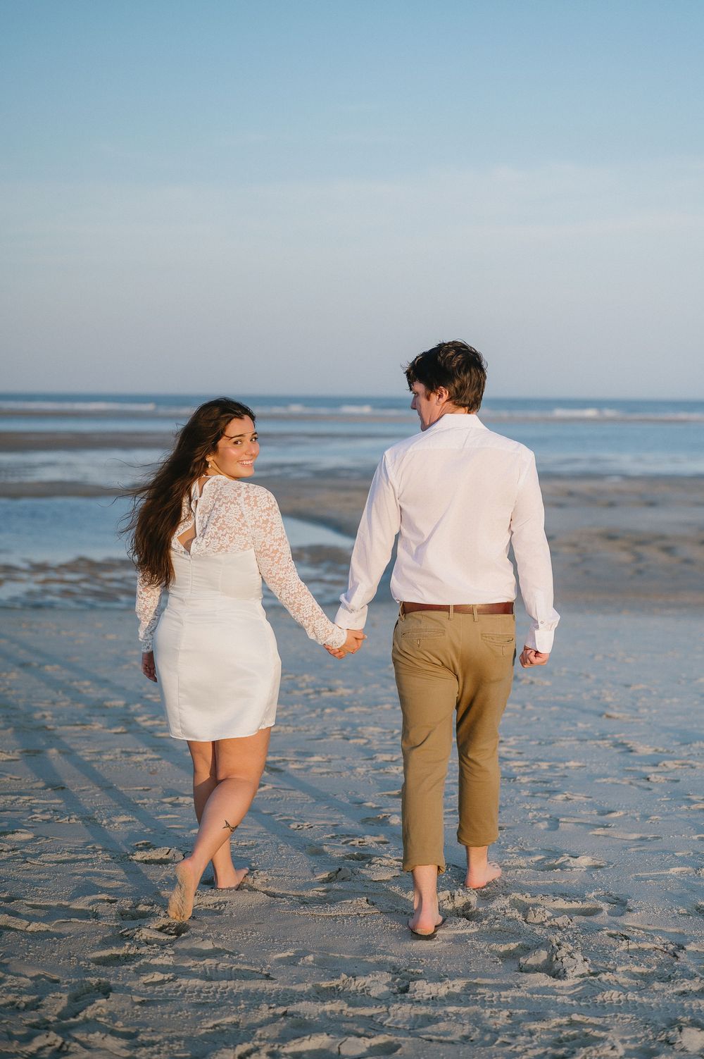 Couple walking barefoot together along beach shoreline during sunset.