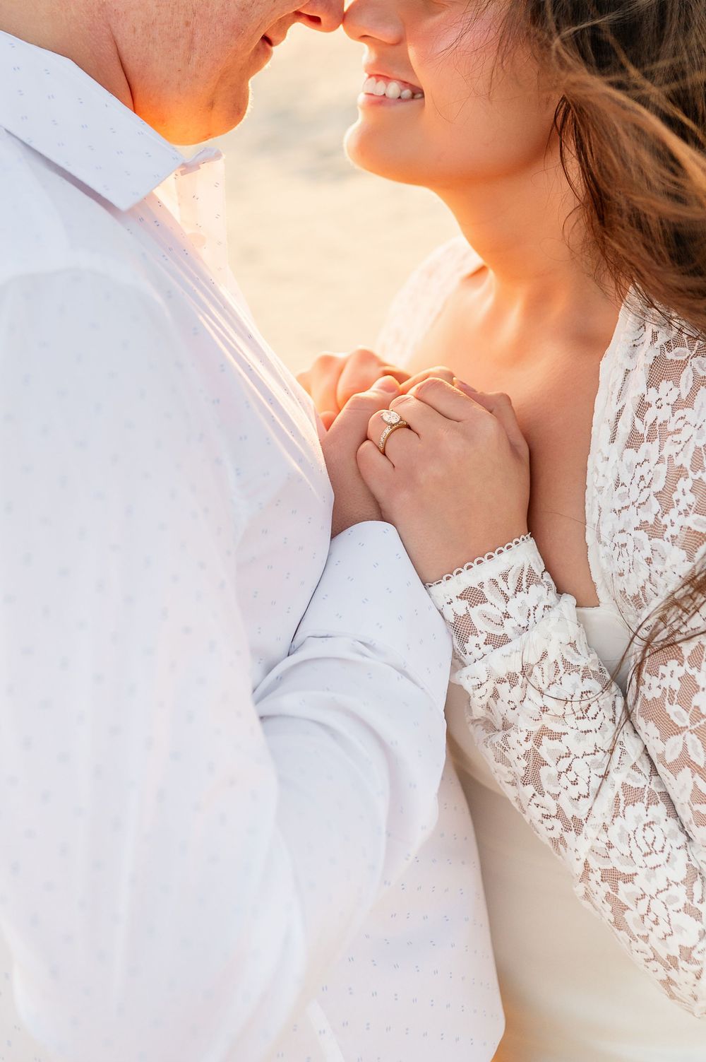 Romantic couple in white embracing at sunset with bride wearing lace sleeves and showing engagement ring.