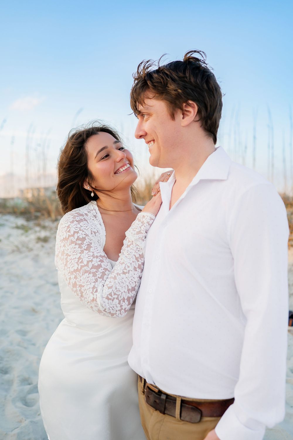 Couple in white clothing share a joyful moment on a sandy beach at sunset.