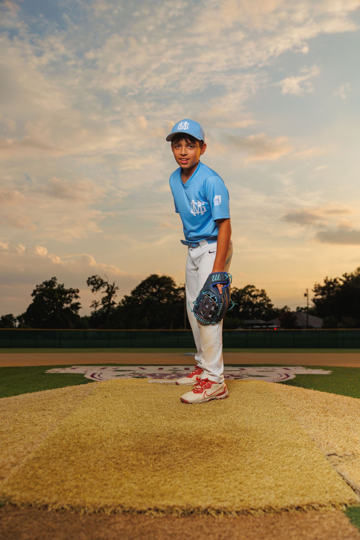 Baseball player stands on pitcher's mound at sunset wearing light blue uniform and red cleats.