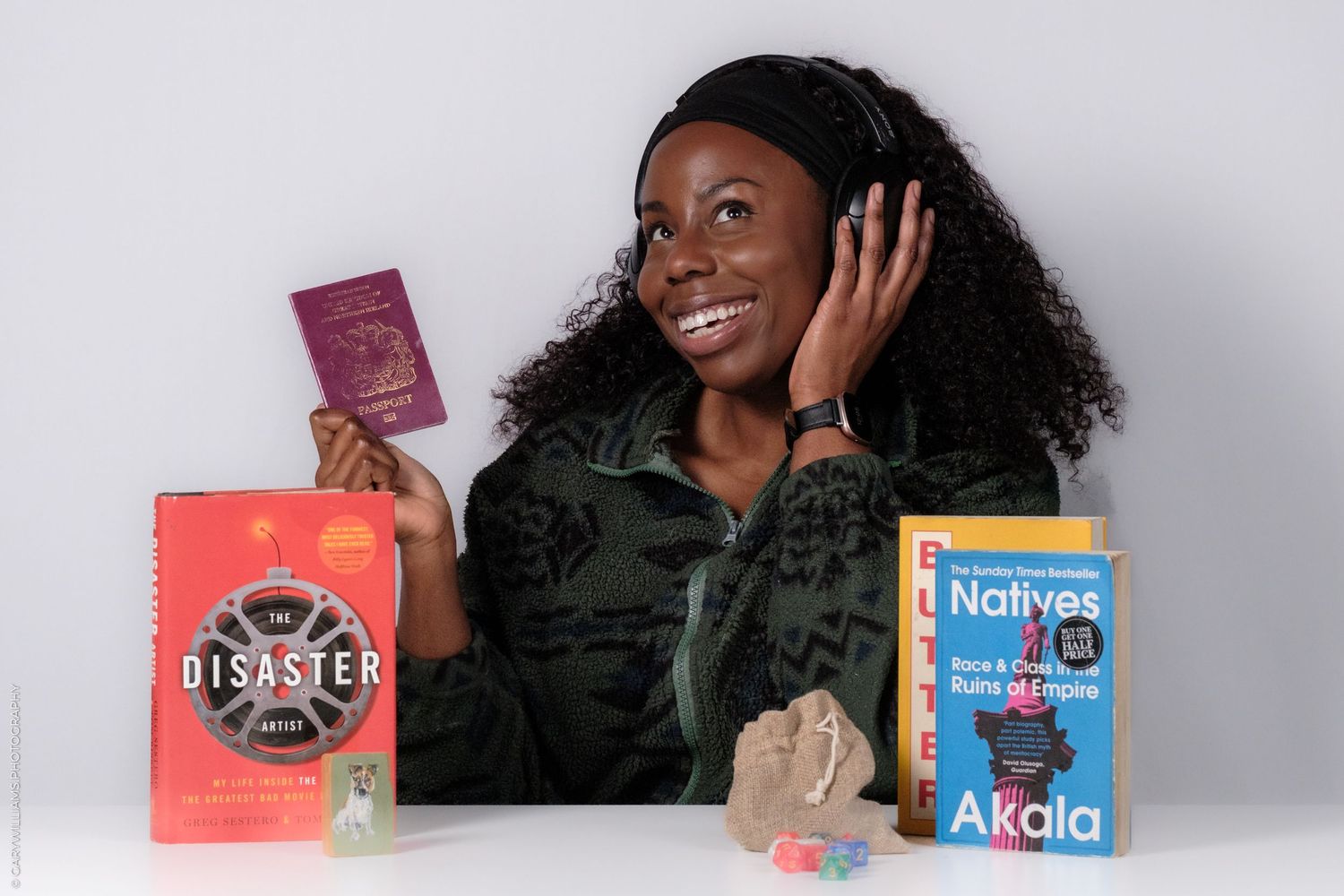 A person wearing headphones holds up a passport while posing with books and other travel items against a gray backdrop.