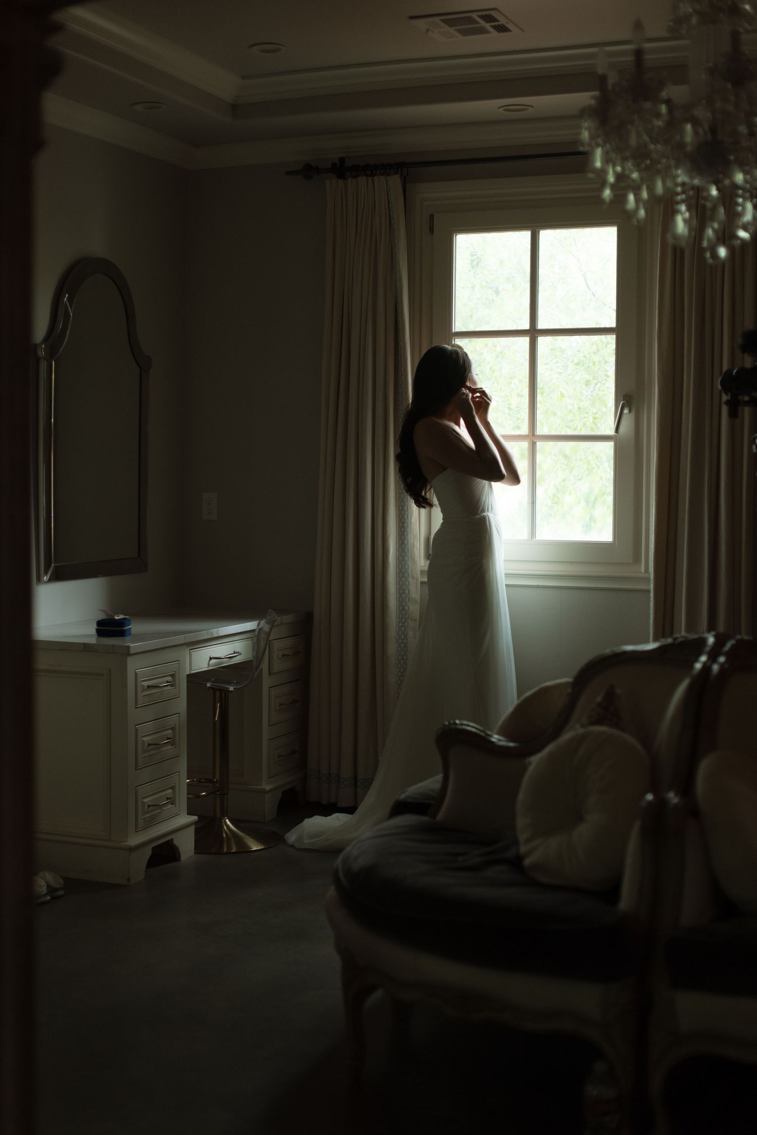 A silhouetted figure in white stands by a window in a moody, dimly lit bedroom with vintage furniture.