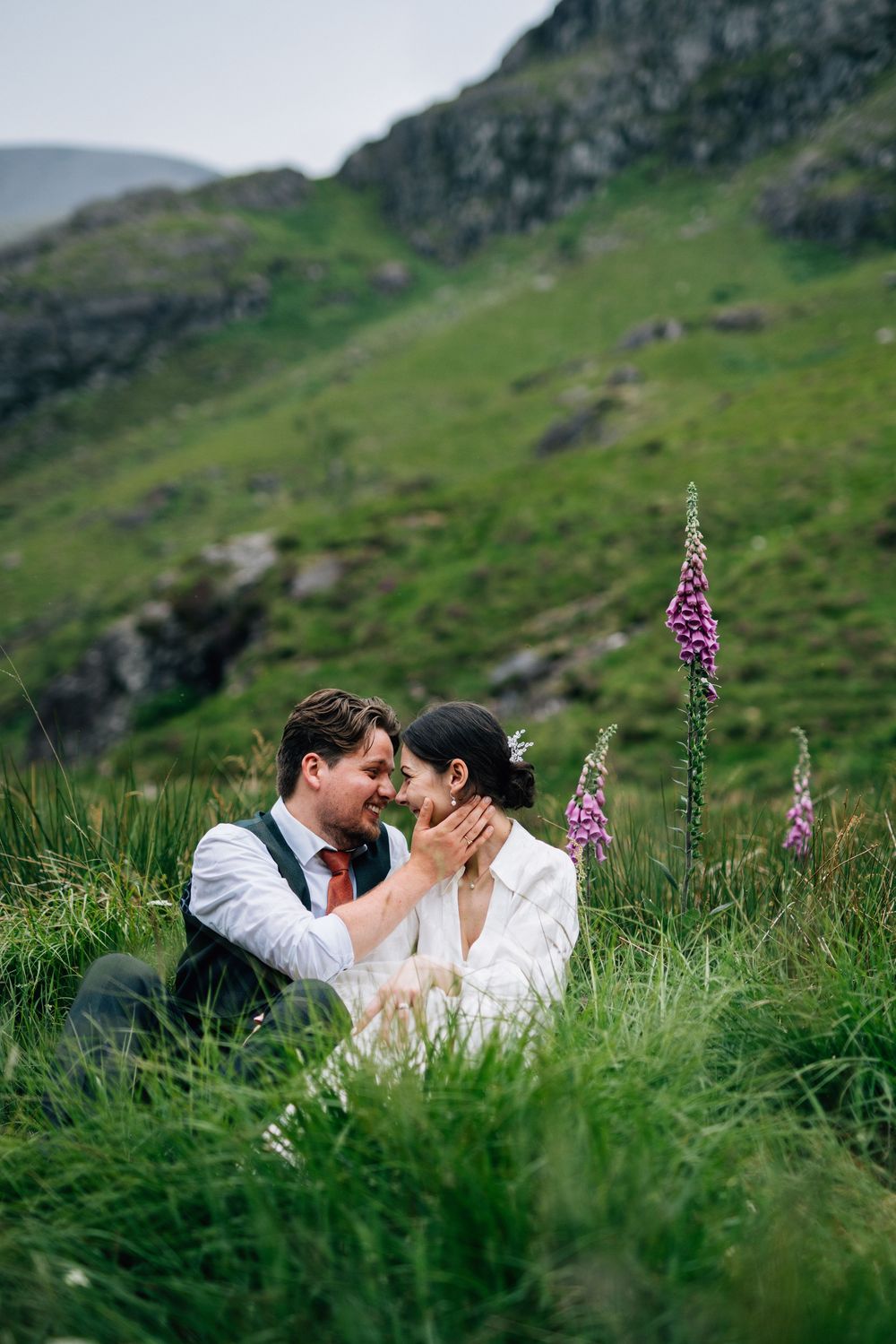 A couple shares an intimate moment surrounded by lush green mountain scenery and wildflowers.