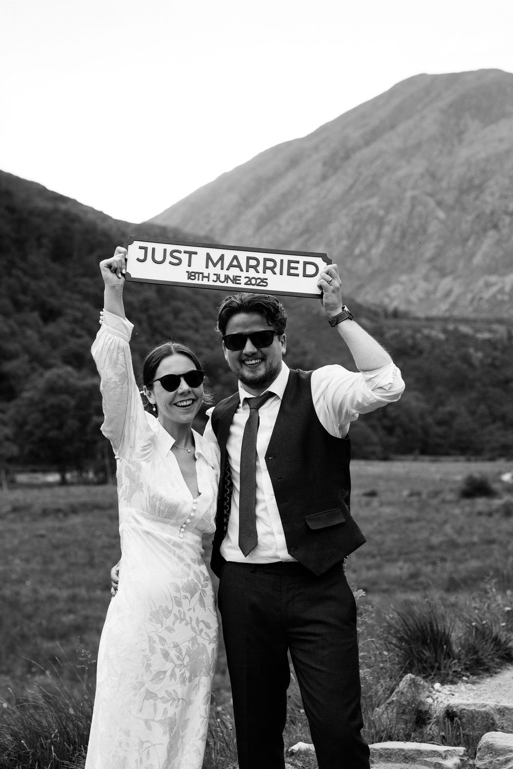 Newlywed couple in sunglasses pose with 'Just Married' sign against mountainous backdrop.