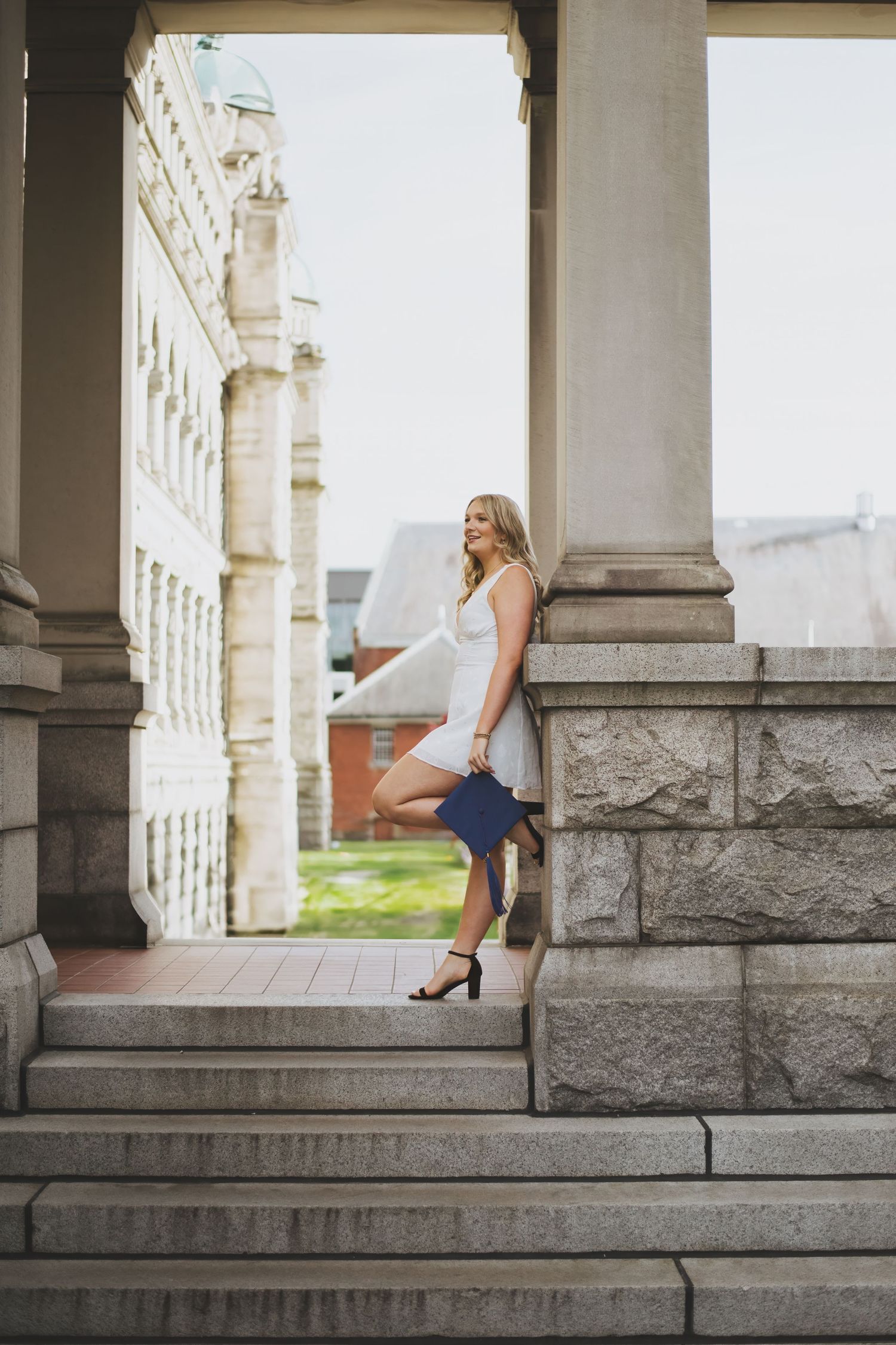 A woman in a white dress sits elegantly on stone steps between classical columns in an architectural setting.