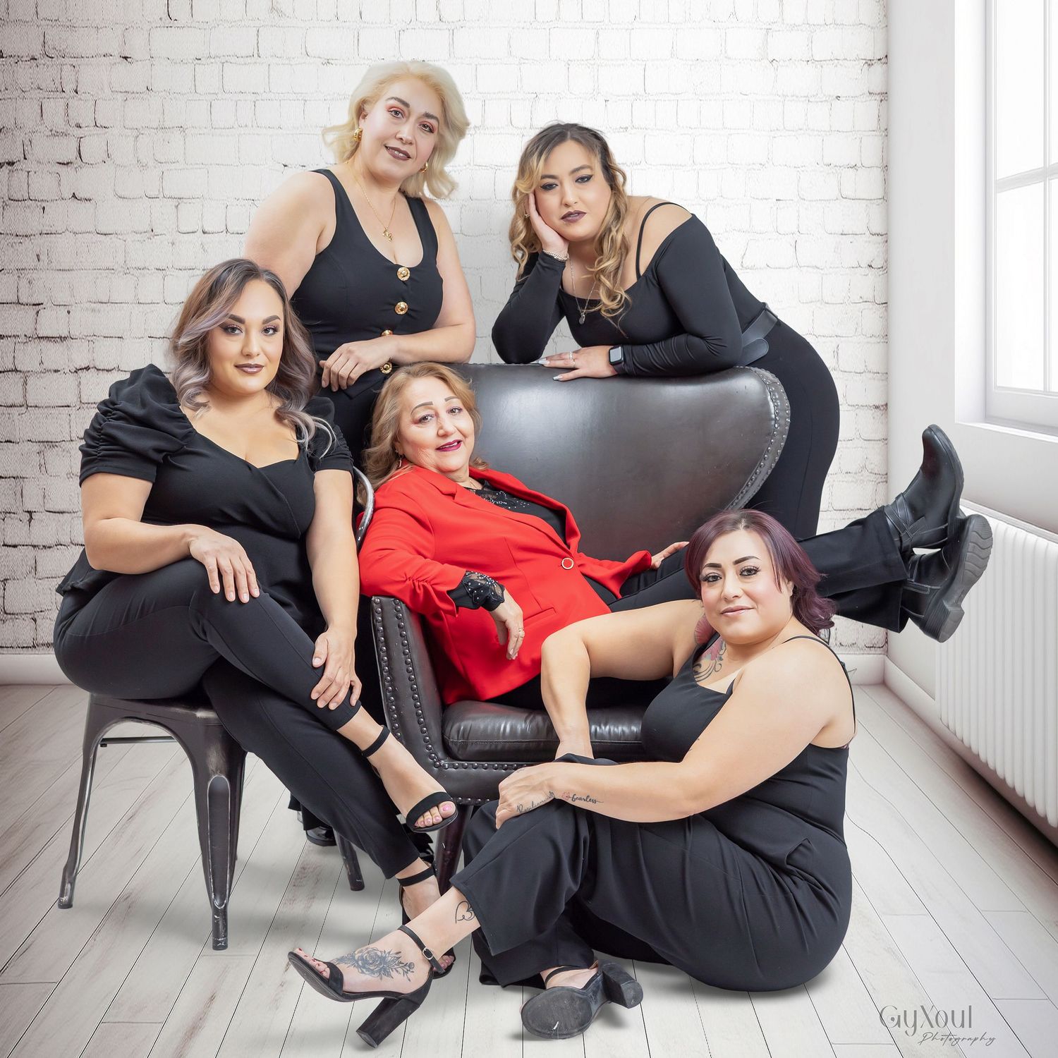 Group in black outfits and red dress posing together on white floor against brick wall in studio setting.