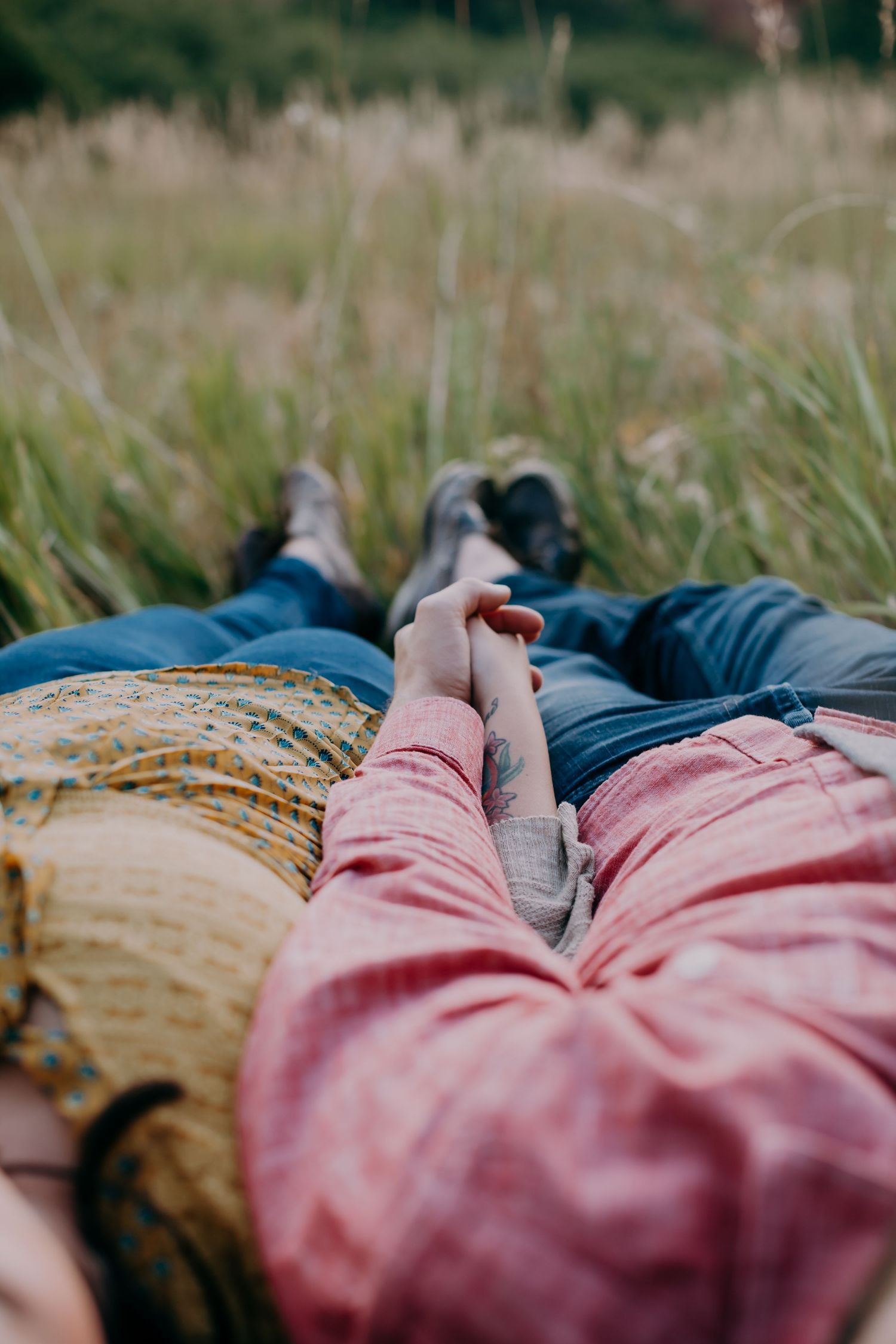 Couple lying in tall grass holding hands, wearing casual jeans and colorful shirts in nature.