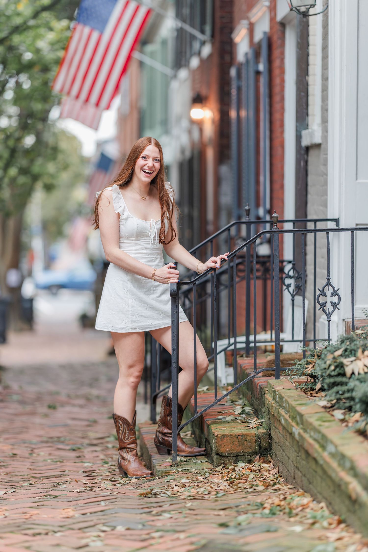 A white summer dress and cowboy boots outfit on a charming brick sidewalk with American flag in the background.