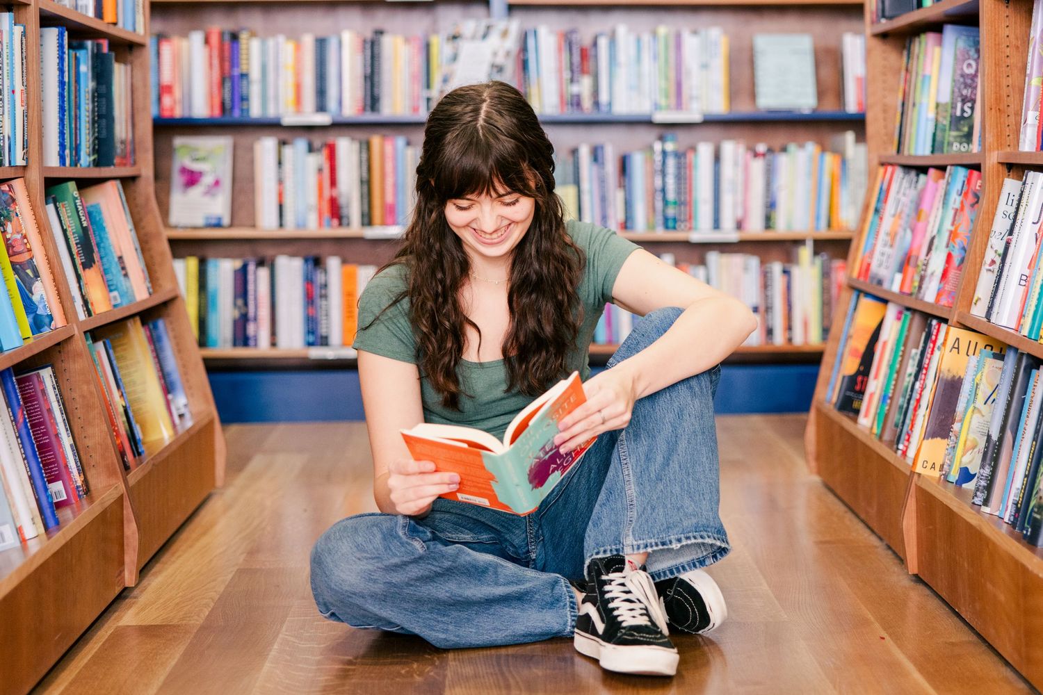 A person sits on the floor reading in a library aisle surrounded by bookshelves.