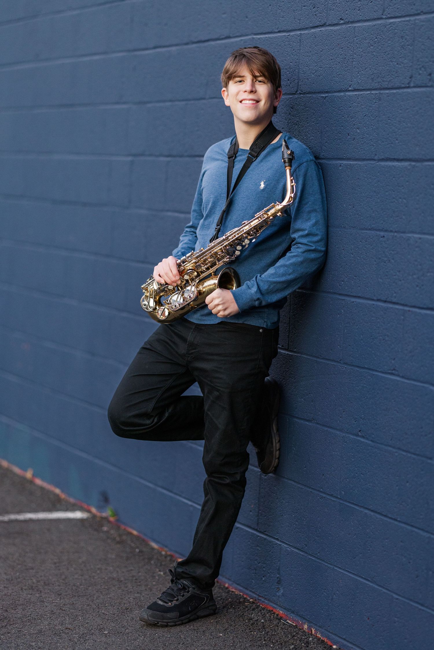 A musician in a blue jacket and black pants poses with a saxophone against a dark blue wall.