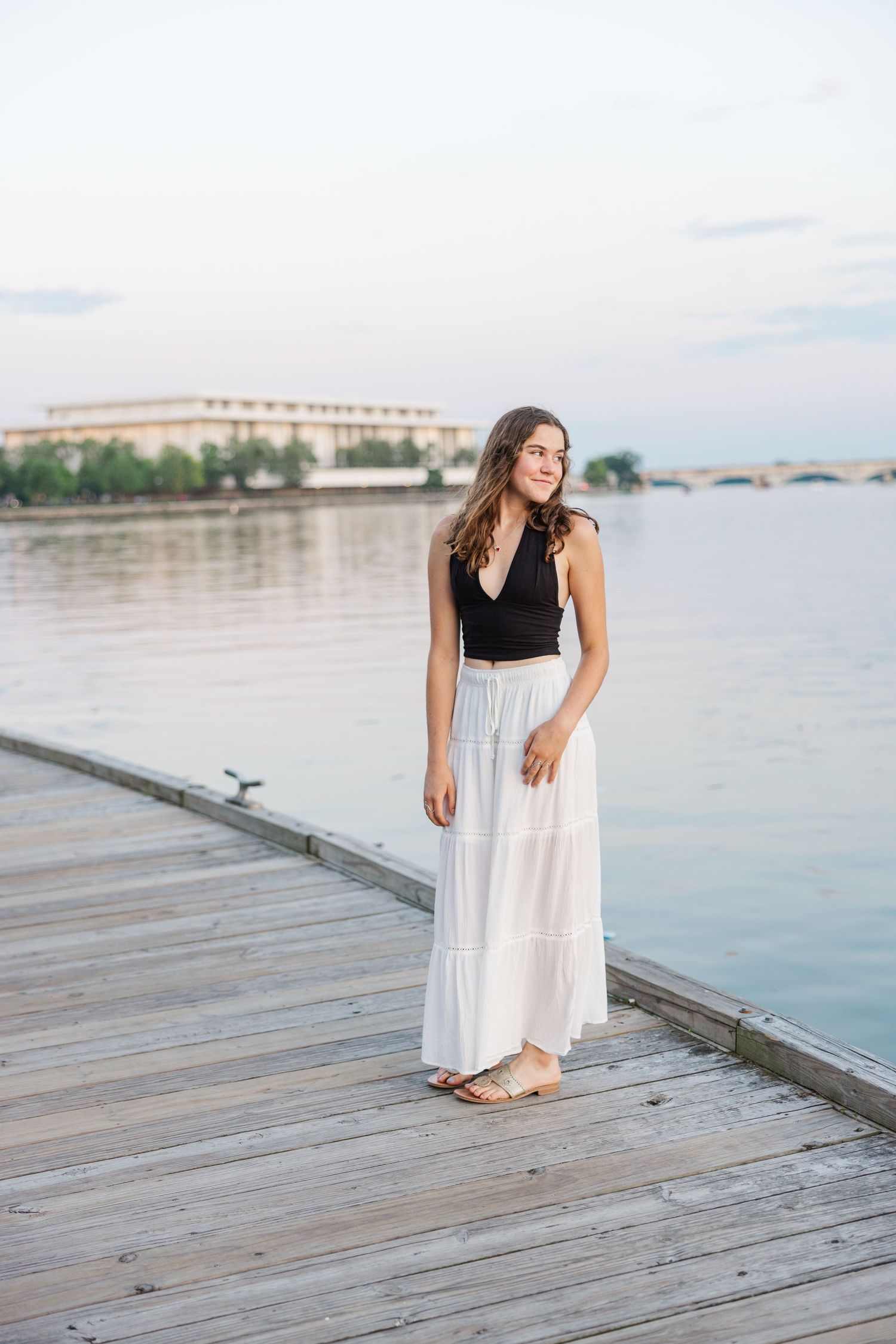 A person in a black top and white maxi skirt poses on a wooden dock by calm waterfront during sunset.