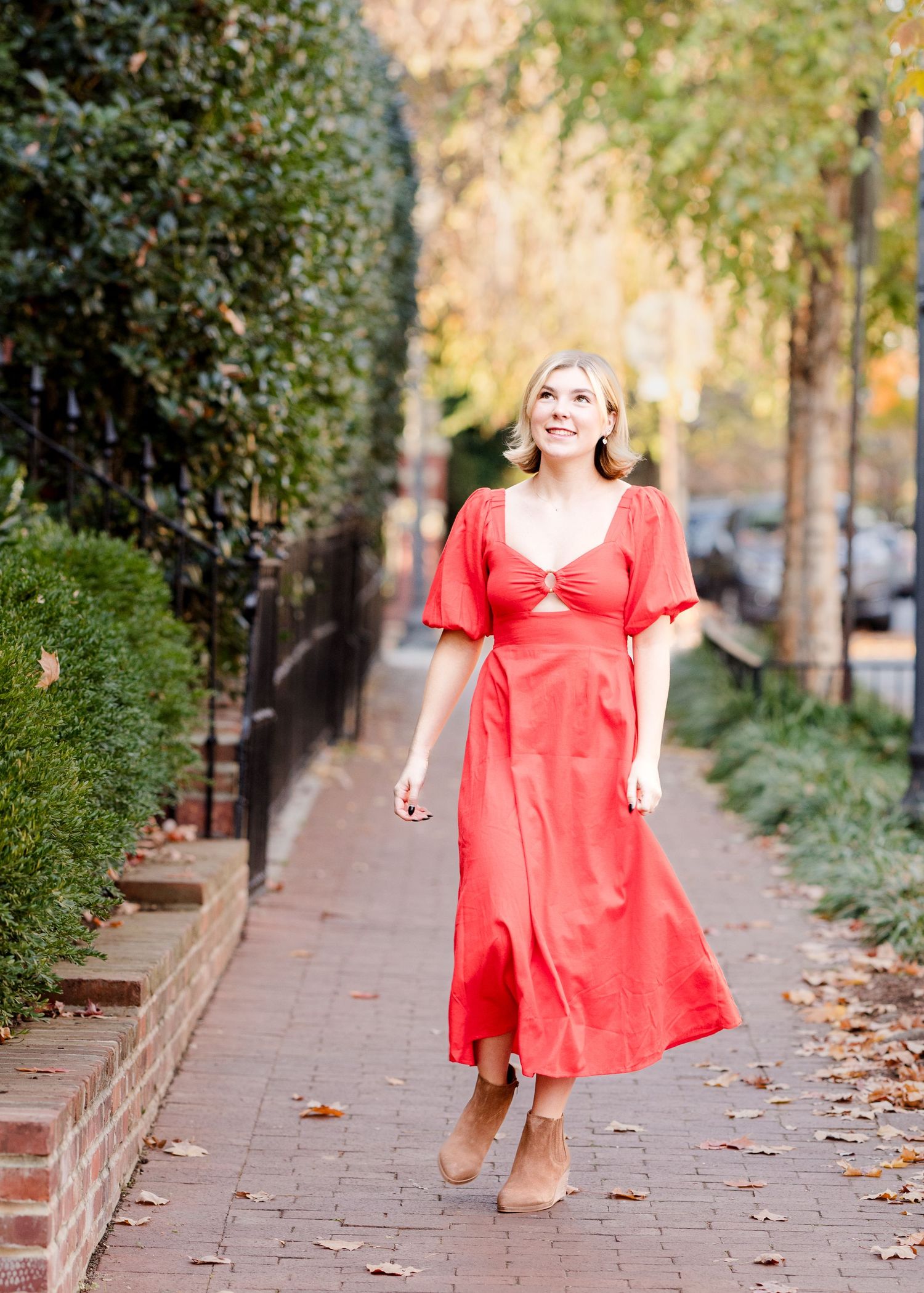 A figure in a flowing coral-colored dress twirls playfully along a tree-lined urban sidewalk on a sunny autumn day.