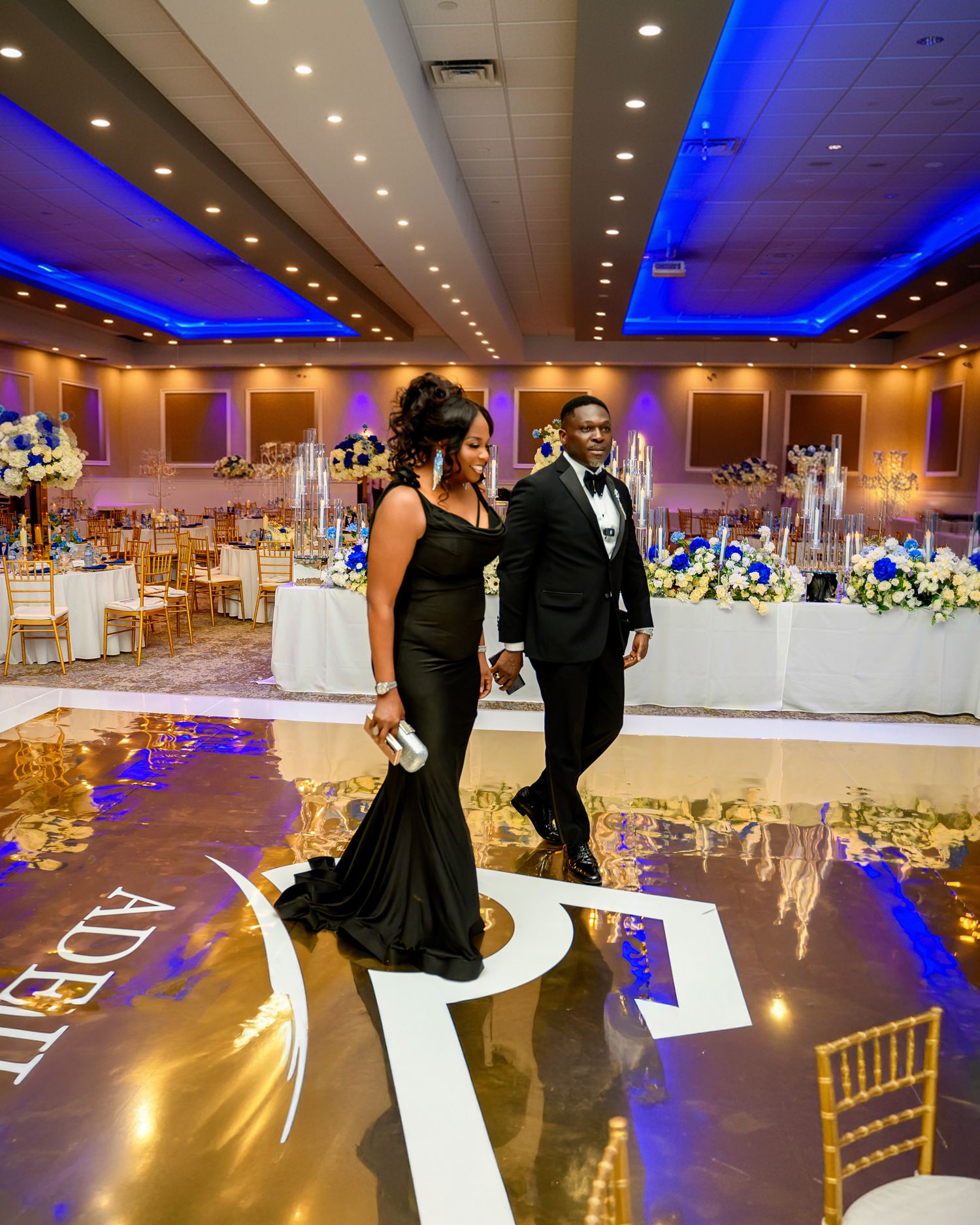 A ballroom decorated with blue uplighting and elegant table settings features guests dancing on a reflective dance floor.