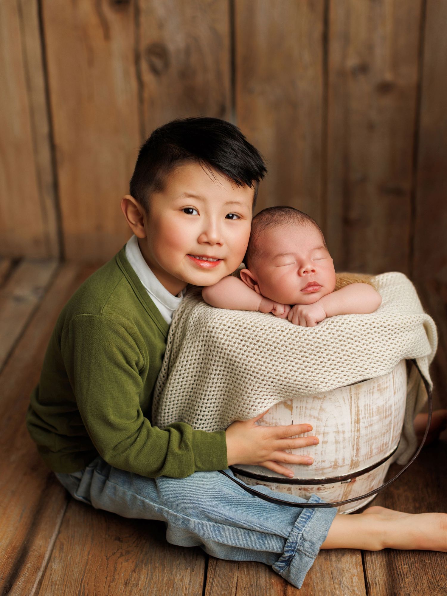 Two children sit together on a wooden floor with a baby in a cream-colored basket against a rustic wood panel backdrop.