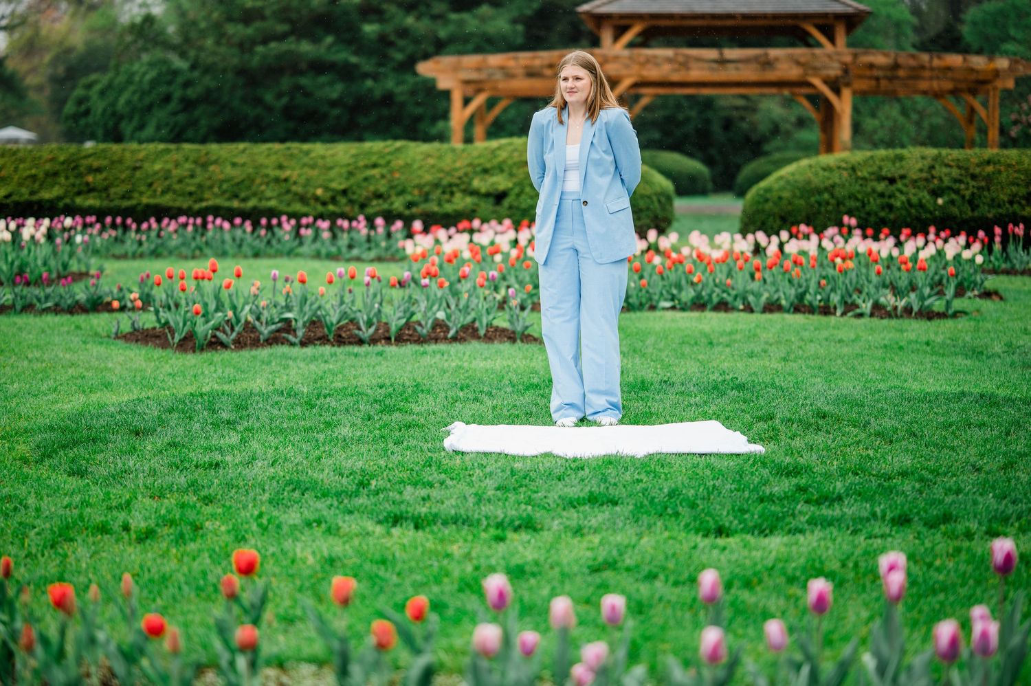 Someone in a light blue outfit stands on a white mat surrounded by colorful tulips in a peaceful garden setting.