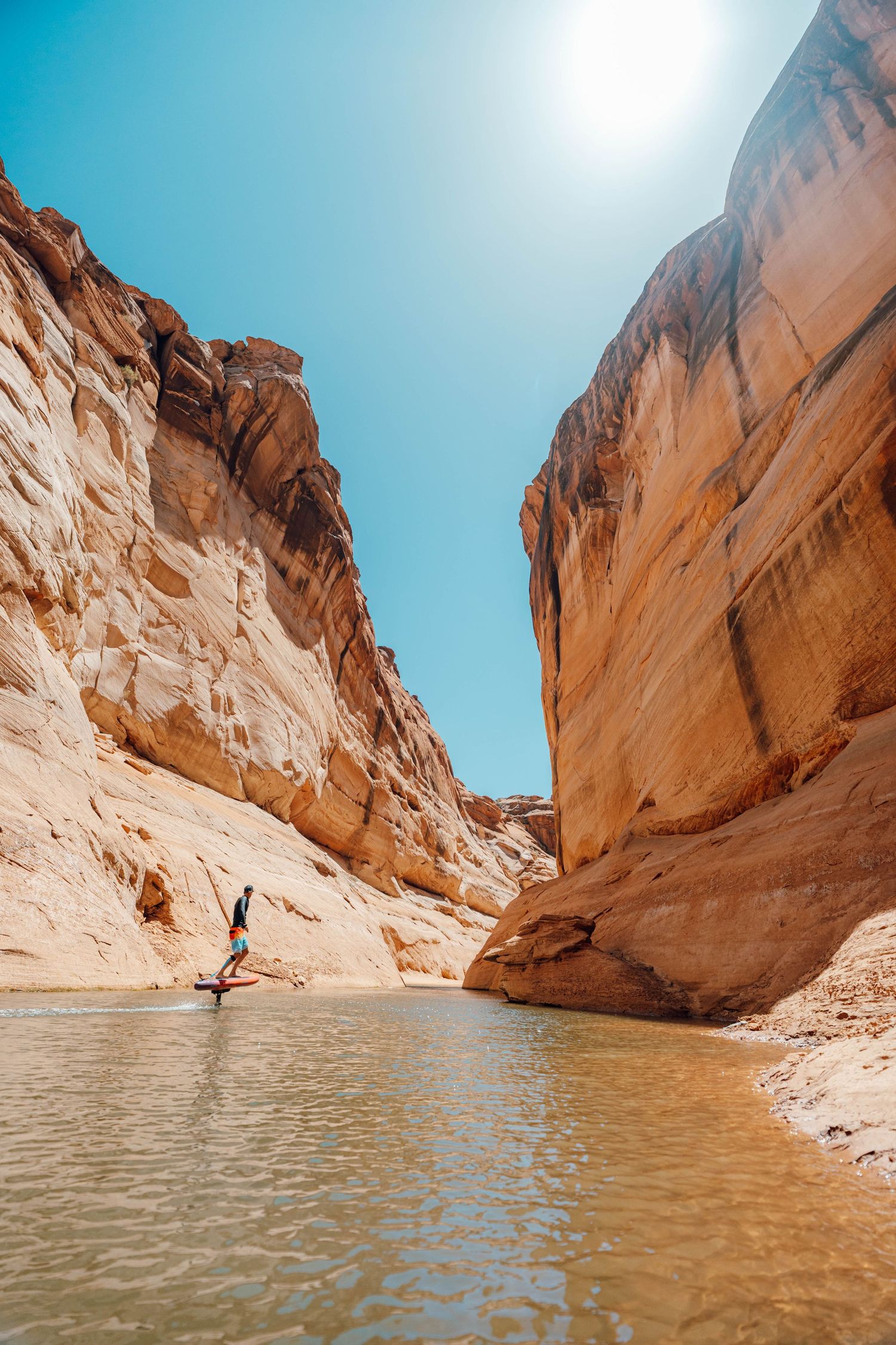 Scenic view of a person hiking through a narrow sandstone canyon with turquoise water and towering orange cliffs.