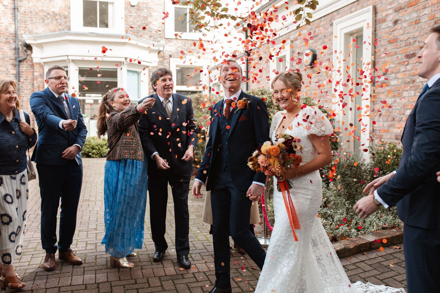 Wedding guests shower the newlyweds with red autumn leaves and petals in front of a rustic brick building.