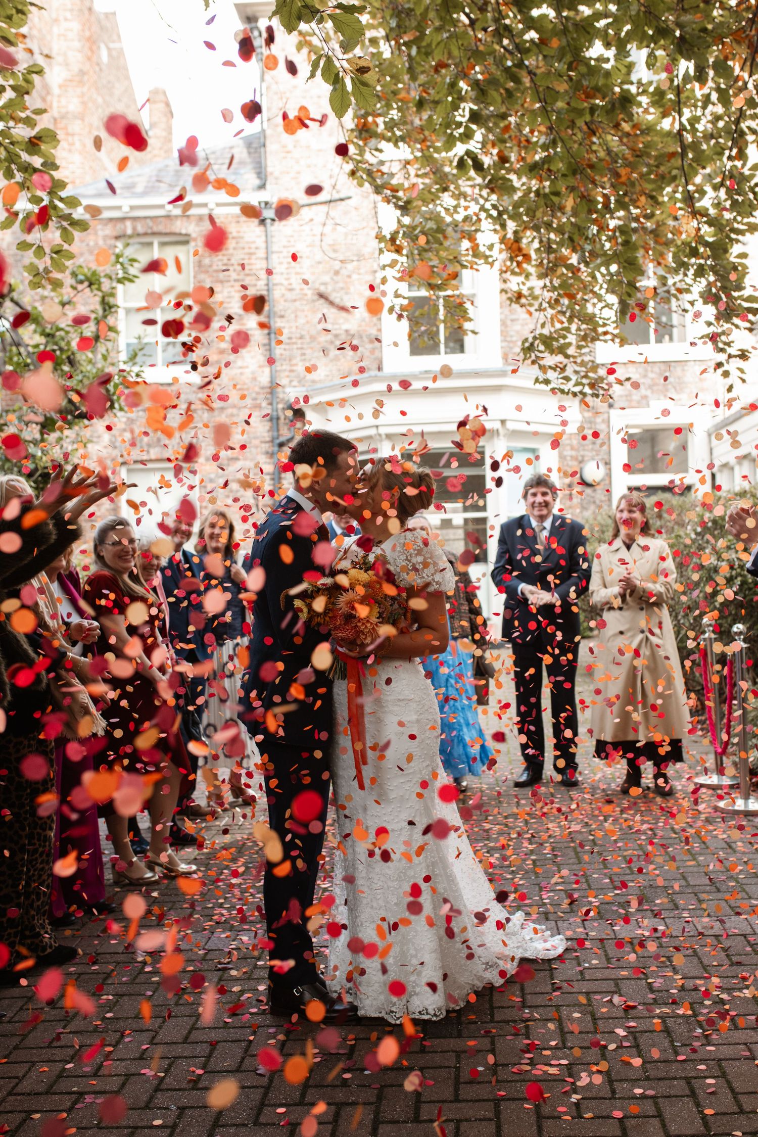 A joyful wedding celebration with red and pink rose petals being tossed in the air on a cobblestone street.