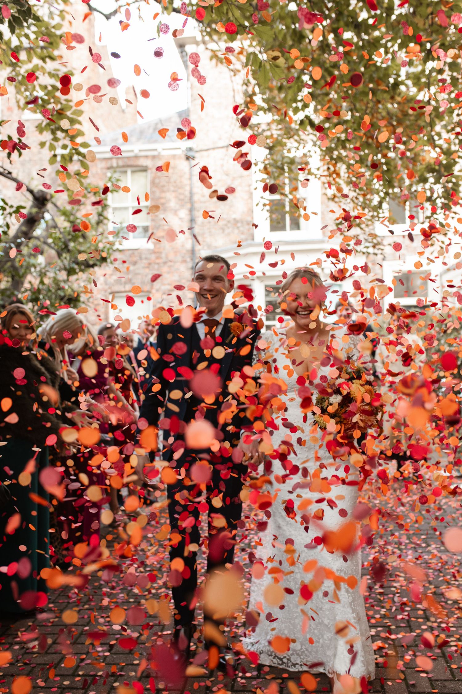 Red rose petals shower down on a couple during an outdoor wedding celebration with a stone building in the background.
