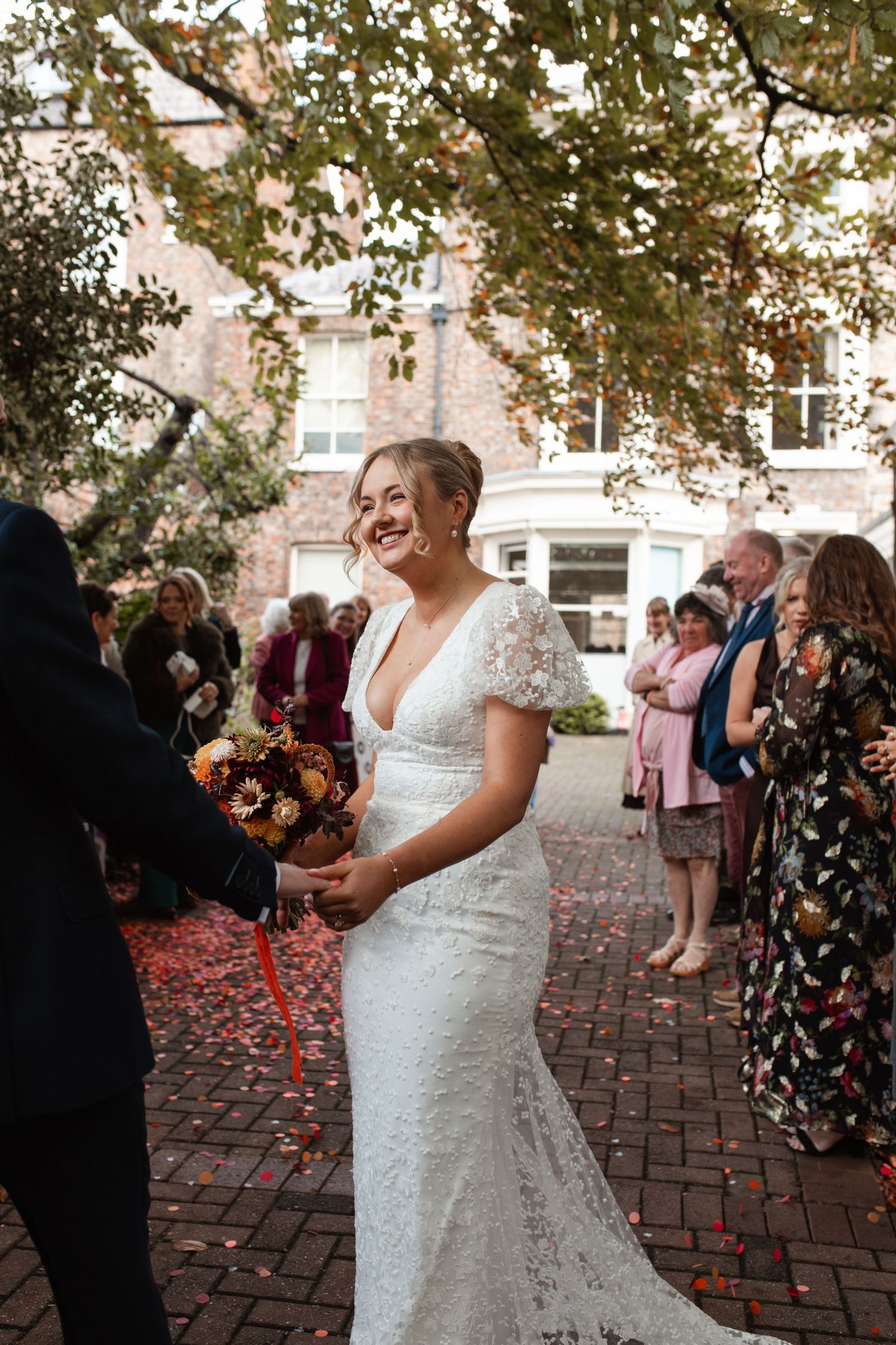 Wedding guests shower confetti on a bride in a white lace dress during an outdoor autumn celebration.