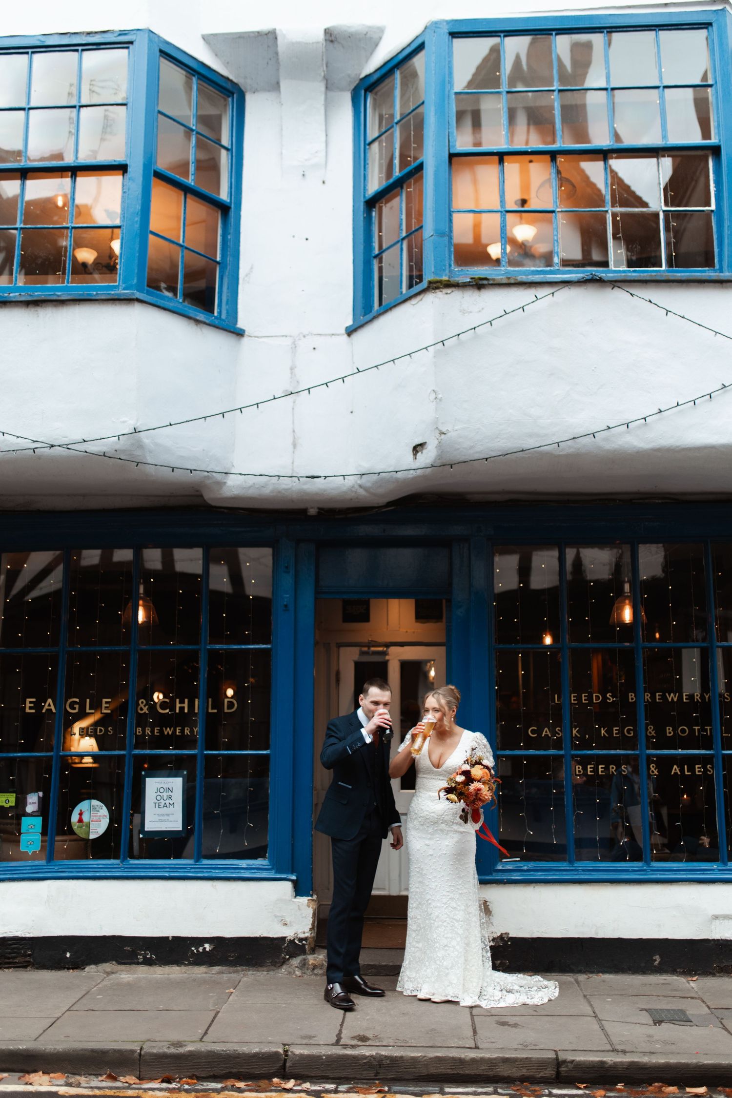 A wedding couple stands in front of a blue-trimmed storefront with large windows and string lights.