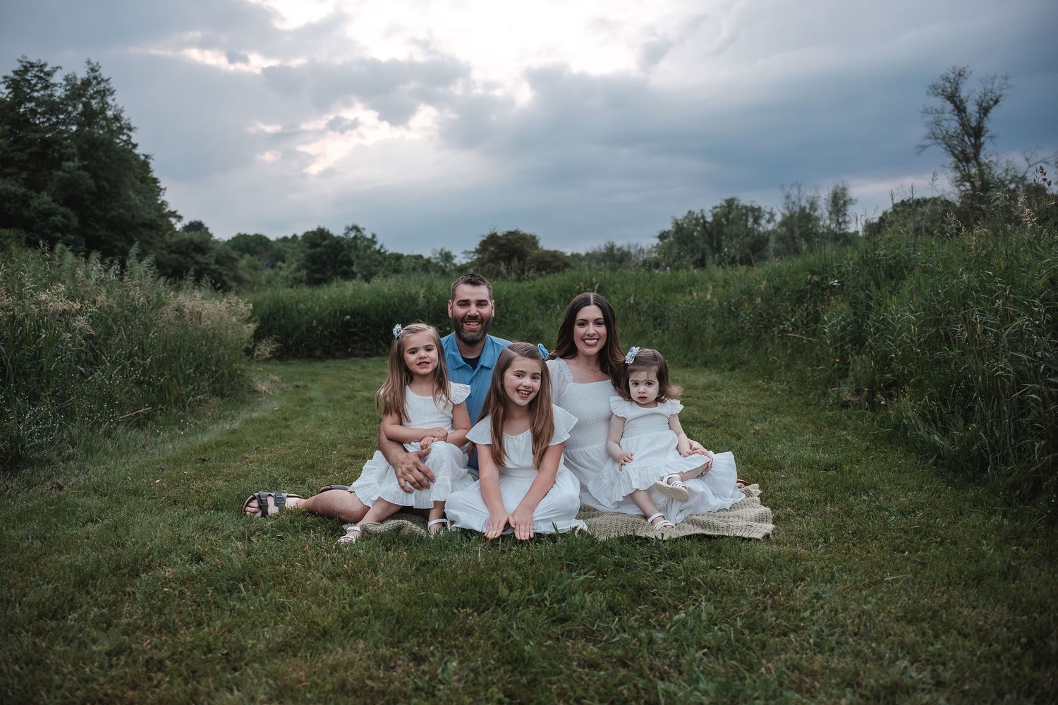 A family sits together on a blanket in a grassy meadow at dusk with dramatic cloudy skies overhead.
