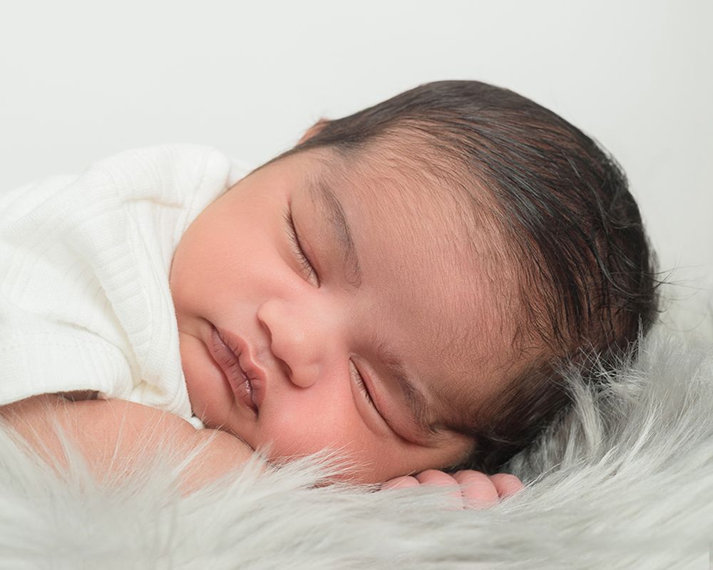 Close-up of a newborn baby with dark hair peacefully sleeping on a soft, grey, fuzzy blanket against a light background