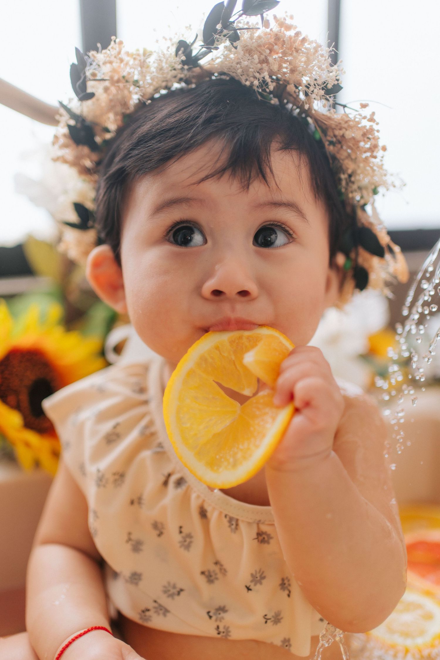A baby wearing a floral crown bites into a fresh orange slice among sunflowers and citrus fruits.