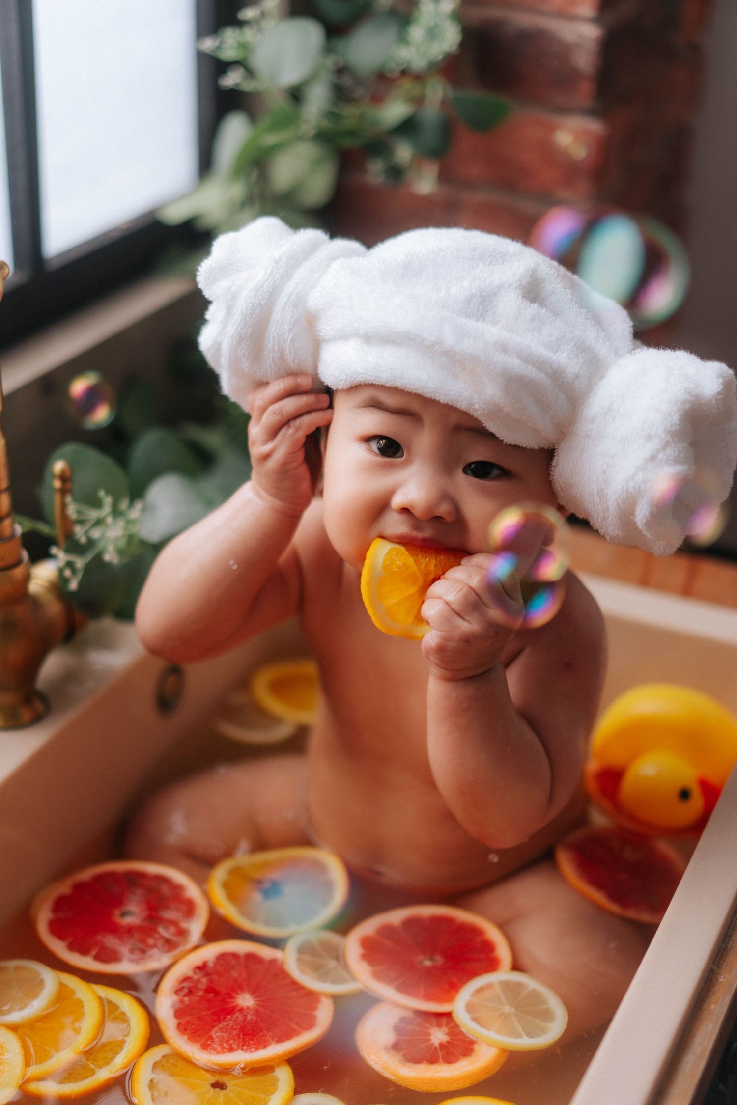 A baby wearing a white towel plays with colorful citrus slices arranged on a white surface.
