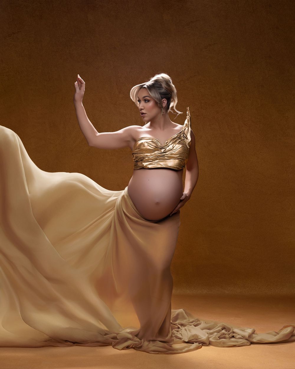 Elegant maternity portrait featuring a golden crop top and flowing champagne-colored gown against a brown studio backdrop.