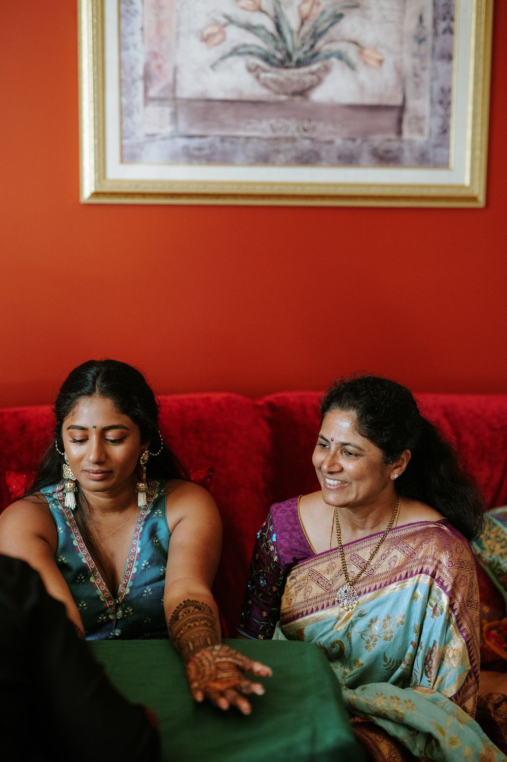 Two friends sharing a joyful moment together on a red couch with traditional Indian attire and jewelry.