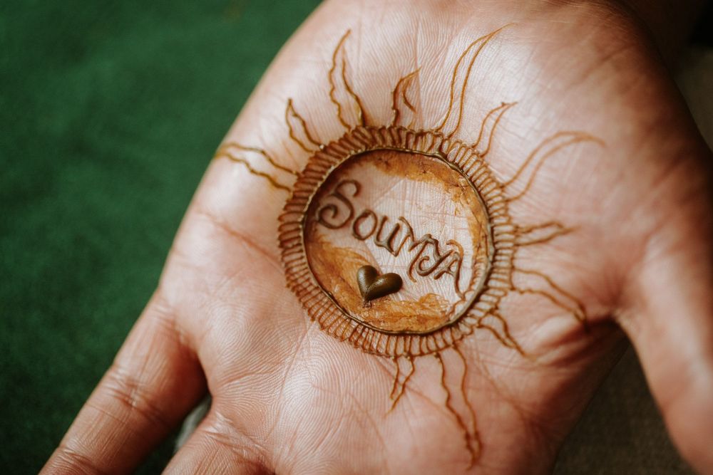 Intricate sun design mehndi henna tattoo drawn on palm of hand against dark green background.