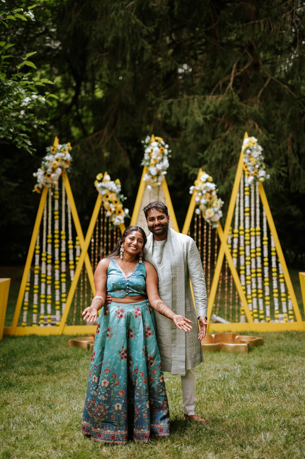 Couple in traditional Indian attire posing in front of yellow decorative backdrop with floral elements.