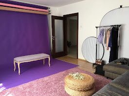 Modern living room with purple carpet, woven ottoman, gray couch, and clothing rack against white walls.