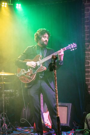 Musician performs on stage with guitar under dramatic green and yellow lighting at live concert venue.